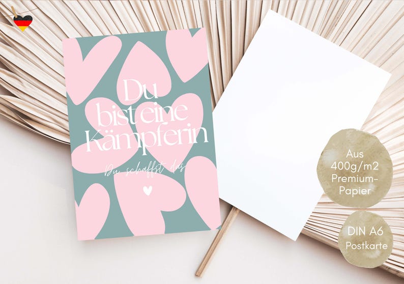 Pu&ograve; includere: Una cartolina con sfondo turchese e forme di cuore rosa. Il testo "Du bist eine K&auml;mpferin" &egrave; visualizzato in bianco. Accanto c'&egrave; una cartolina bianca vuota. La cartolina &egrave; realizzata in carta premium da 400 g/m2.
