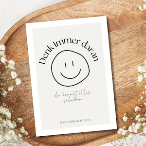May include: A rectangular card with a light beige background featuring a black smiley face and text in German. The card is on a wooden tray, surrounded by white flowers.