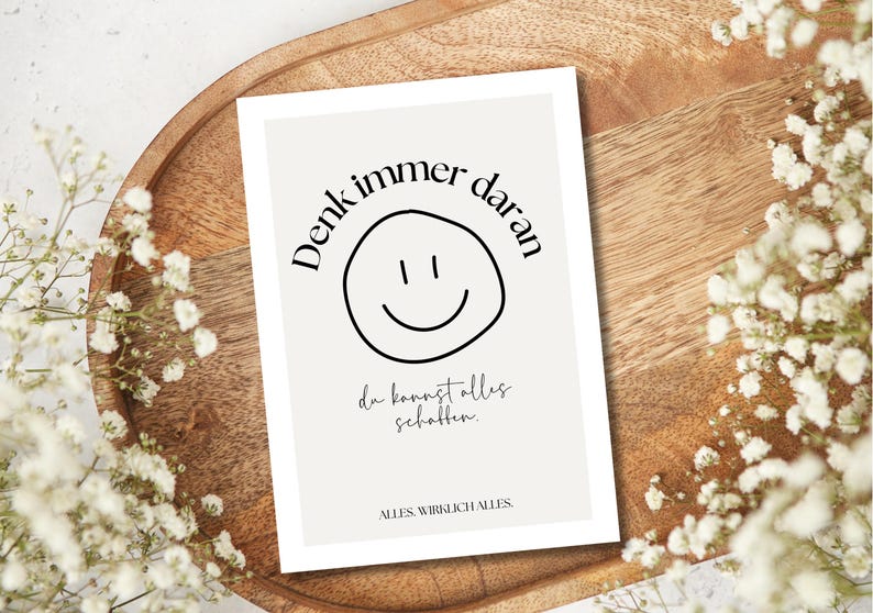 May include: A rectangular card with a light beige background featuring a black smiley face and text in German. The card is on a wooden tray, surrounded by white flowers.