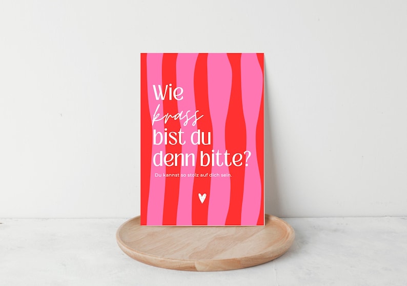 May include: A rectangular greeting card featuring a vertical wavy striped pattern in alternating shades of bright red and hot pink. The card displays the white text "Wie krass bist du denn bitte?" followed by "Du kannst so stolz auf dich sein." and a small white heart icon.