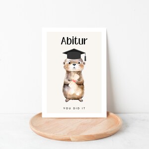 May include: A graduation card with a cartoon otter wearing a black graduation cap and holding a diploma. The card has the text "Abitur" and "You Did It" on it.