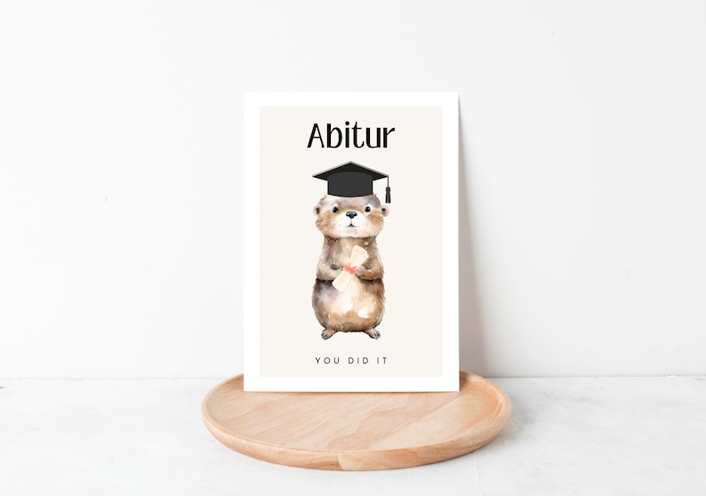 May include: A graduation card with a cartoon otter wearing a black graduation cap and holding a diploma. The card has the text "Abitur" and "You Did It" on it.
