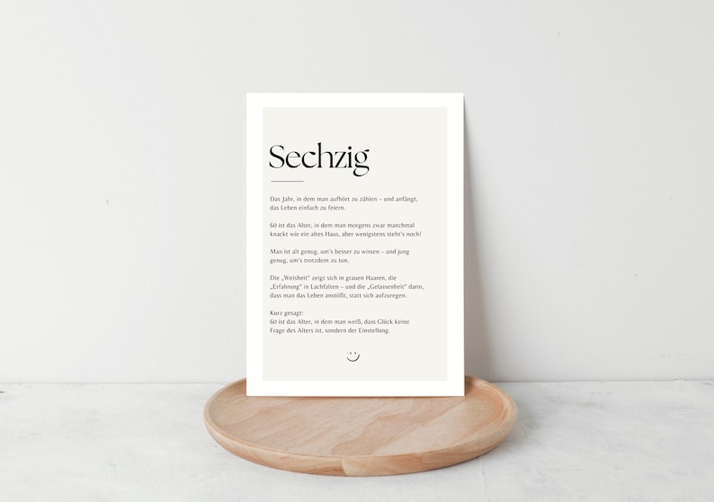 May include: A framed print with the German word "Sechzig" at the top, meaning sixty. The print has a cream-colored background with black text. It is displayed on a round, light brown wooden tray.