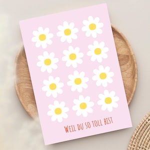 Könnte beinhalten: Eine rosa Grußkarte mit einem Muster aus weißen Gänseblümchen mit gelben Zentren. Die Karte trägt den deutschen Text "WEIL DU SO TOLL BIST" in Rot. Die Karte wird auf einem Holztablett präsentiert.