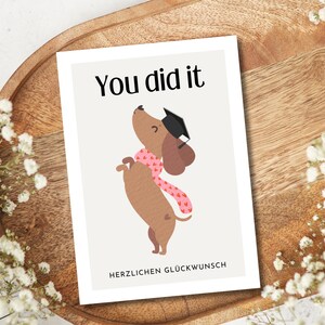 May include: A graduation card with a cartoon dachshund wearing a graduation cap and a pink scarf. The card reads "You did it" at the top and "Herzlichen Gl&uuml;ckwunsch" at the bottom. The card is on a wooden tray with white flowers.