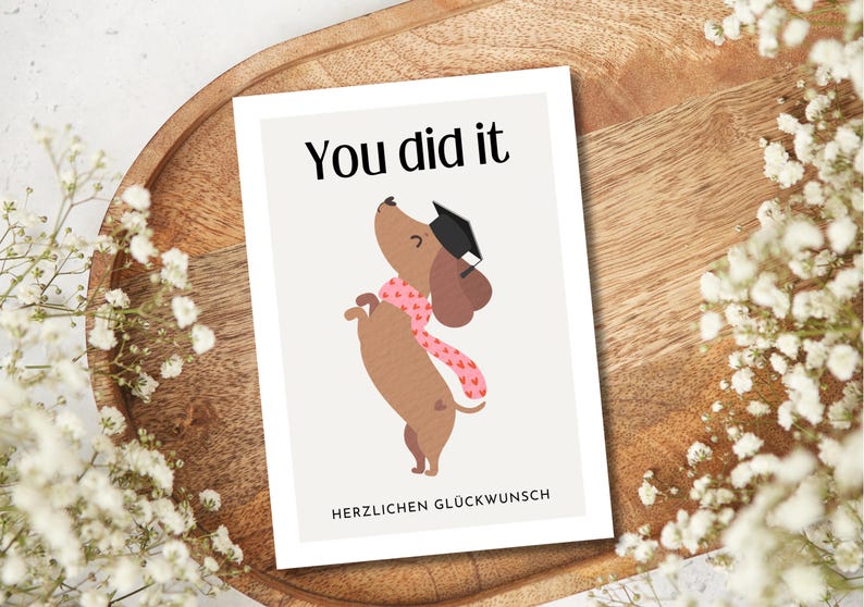 May include: A graduation card with a cartoon dachshund wearing a graduation cap and a pink scarf. The card reads "You did it" at the top and "Herzlichen Gl&uuml;ckwunsch" at the bottom. The card is on a wooden tray with white flowers.