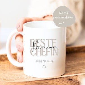 May include: White ceramic mug with a black inscription that reads "Beste Marina Chefin" and "Danke fur alles" with a smiley face. The mug has a pink handle.