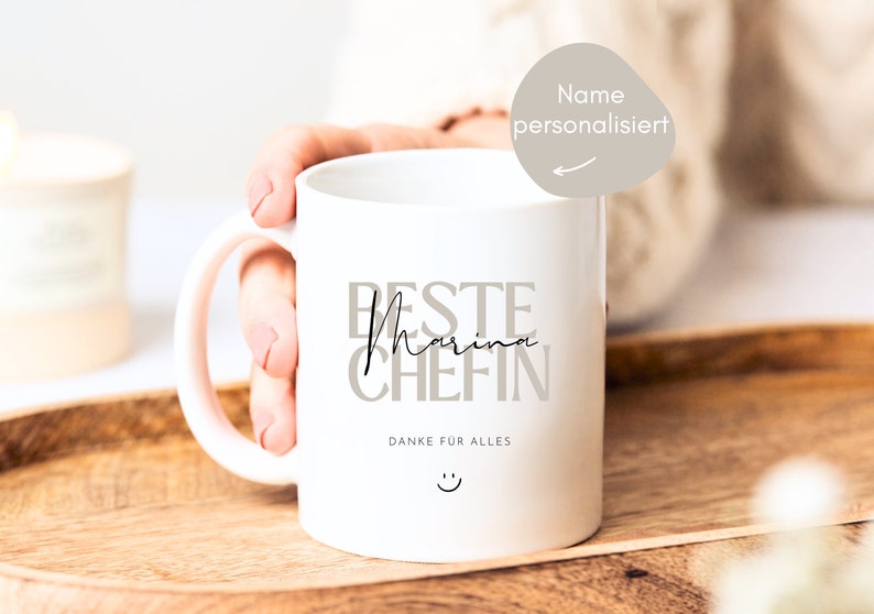 May include: White ceramic mug with a black inscription that reads "Beste Marina Chefin" and "Danke fur alles" with a smiley face. The mug has a pink handle.