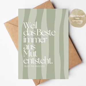 May include: A rectangular greeting card with a sage green background featuring vertical wavy lines and white text that reads "Weil das Beste immer aus Mut entsteht. TRUST THE PROCESS." The card is placed on top of a brown paper envelope.