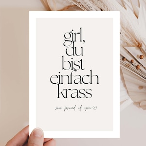Peut inclure: Impression rectangulaire avec les mots "girl, du bist einfach krass" en script noir. En dessous, le texte "sooo proud of you" avec un symbole de cœur. L'impression est tenue par une main, avec des plantes séchées en arrière-plan.