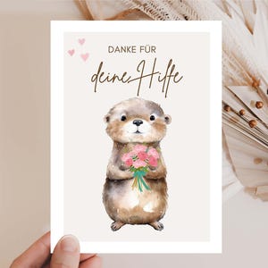 Peut inclure: Carte de vœux avec une illustration à l'aquarelle d'une loutre tenant un bouquet de fleurs roses. La carte porte l'inscription "DANKE FÜR deine Hilfe" et trois petits cœurs roses. La loutre est marron et beige avec un ventre blanc.
