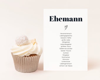 May include: A white card with black text that reads "Ehemann" with a heart symbol and a list of words describing a husband. The card is next to a white cupcake with white frosting and a white coconut ball on top.