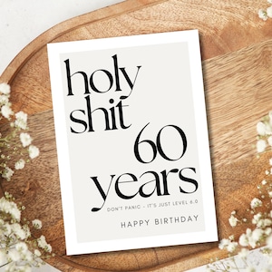 May include: A white card with black text that reads "holy shit 60 years." The card is on a wooden tray, surrounded by white flowers. The text "DON'T PANIC - IT'S JUST LEVEL 6.0" and "HAPPY BIRTHDAY" are also on the card.