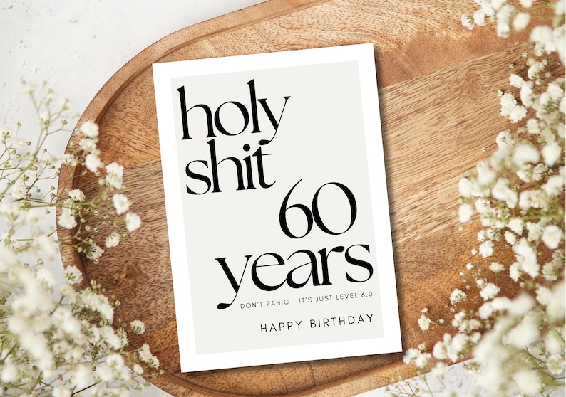 May include: A white card with black text that reads "holy shit 60 years." The card is on a wooden tray, surrounded by white flowers. The text "DON'T PANIC - IT'S JUST LEVEL 6.0" and "HAPPY BIRTHDAY" are also on the card.