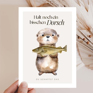 Könnte beinhalten: Eine Grußkarte mit einer Aquarellillustration eines Otters, der einen Fisch hält. Die Karte enthält deutschen Text mit der Aufschrift "Halt noch ein bisschen Dorsch" und "DU SCHAFFST DAS". Der Hintergrund ist hellbeige.