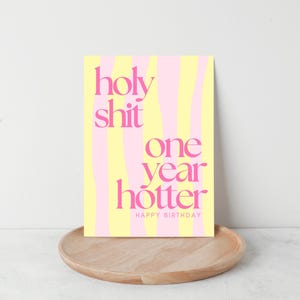 May include: A birthday card with the text "holy shit one year hotter HAPPY BIRTHDAY" in pink, set against a yellow and pink striped background. The card is displayed on a wooden tray.