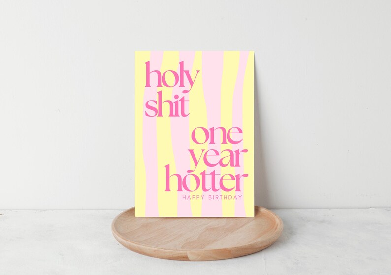 May include: A birthday card with the text "holy shit one year hotter HAPPY BIRTHDAY" in pink, set against a yellow and pink striped background. The card is displayed on a wooden tray.