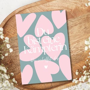 Pu&ograve; includere: Un biglietto di auguri con sfondo verde acqua e forme di cuore rosa. La carta ha il testo tedesco "Du bist eine K&auml;mpferin" in bianco, che significa "Sei una combattente". La carta &egrave; su un vassoio di legno, circondata da fiori bianchi.