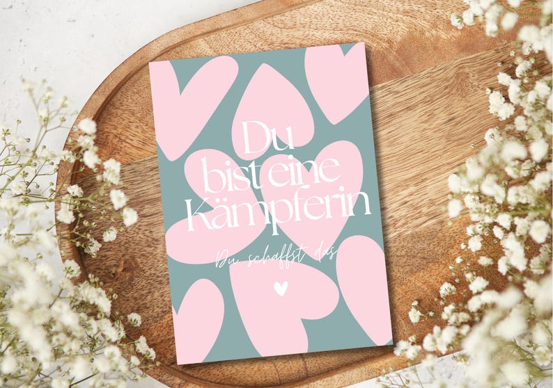 Pu&ograve; includere: Un biglietto di auguri con sfondo verde acqua e forme di cuore rosa. La carta ha il testo tedesco "Du bist eine K&auml;mpferin" in bianco, che significa "Sei una combattente". La carta &egrave; su un vassoio di legno, circondata da fiori bianchi.