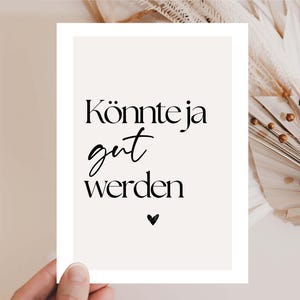 May include: A rectangular print with the German text "Könnte ja gut werden" in black script and a small heart. The print is held by a hand, with a neutral background and decorative dried plants.