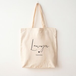 May include: Cream-colored tote bag with the word "Lawyer" in a black script font, a small black heart, and the name "Antonia" below. The bag has long handles and is hanging against a white background.