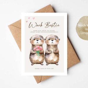 May include: A greeting card with two watercolor otters, one holding flowers. The card reads "Work Bestie" with German text below. A brown envelope is behind the card. A round sticker with German text is on the right.