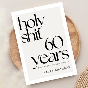 May include: A greeting card with the text "holy shit 60 years" in black font. The card also includes the phrases "DON'T PANIC - IT'S JUST LEVEL 6.0" and "HAPPY BIRTHDAY". The card is on a wooden plate.