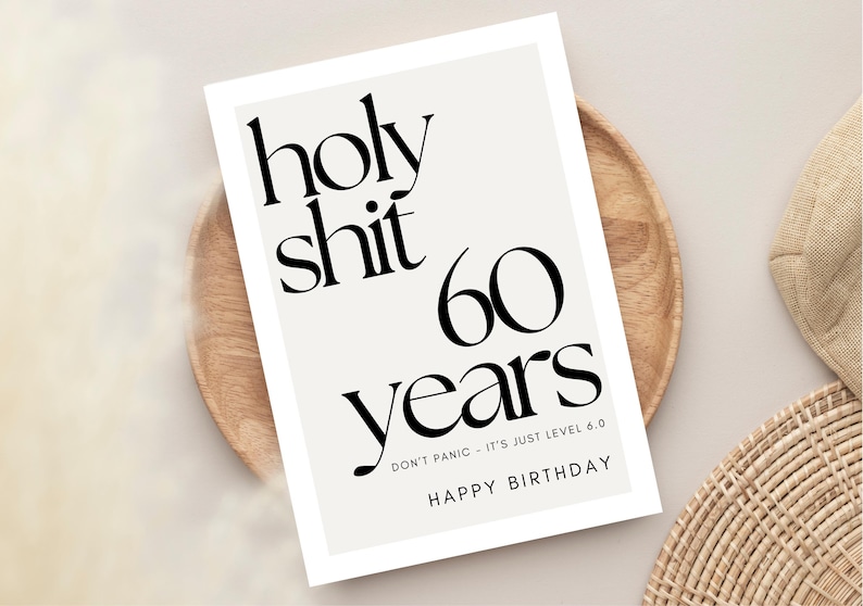 May include: A greeting card with the text "holy shit 60 years" in black font. The card also includes the phrases "DON'T PANIC - IT'S JUST LEVEL 6.0" and "HAPPY BIRTHDAY". The card is on a wooden plate.