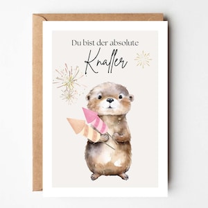 Peut inclure: Carte de vœux avec une illustration à l'aquarelle d'une loutre tenant des fusées jouets. La carte porte le texte allemand "Du bist der absolute Knaller" avec des accents dorés en forme d'éclats d'étoiles. Fond crème.