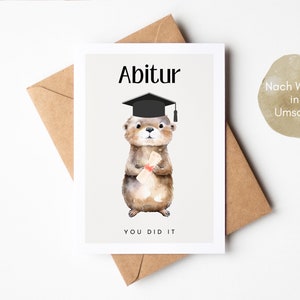 May include: A graduation card featuring a cartoon otter wearing a graduation cap and holding a diploma. The card has the text "Abitur" and "You Did It" on it. The card is on a brown envelope. The text "Nach Wunsch inkl. Umschlag" is on the envelope.