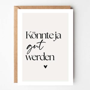 Puede incluir: Una impresión artística rectangular con el texto alemán "Könnte ja gut werden" en escritura negra y un pequeño símbolo de corazón. La impresión está sobre un fondo crema y está en un sobre marrón.
