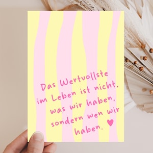 Può includere: Una carta rettangolare con sfondo a righe gialle e rosa. La carta presenta il testo "Das Wertvollste im Leben ist nicht, was wir haben, sondern wen wir haben." in rosa, con un cuore rosa alla fine.