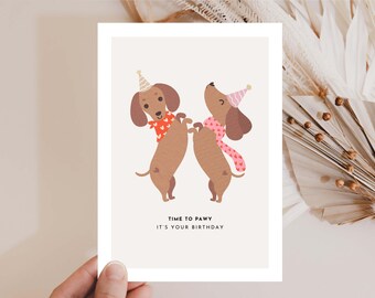 Puede incluir: Tarjeta de felicitación con dos perros salchicha de dibujos animados celebrando un cumpleaños. Los perros marrones llevan gorros de fiesta y bufandas. El texto "TIME TO PAWY IT'S YOUR BIRTHDAY" está en negro sobre fondo crema. Diseño festivo.