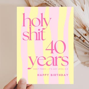 Könnte beinhalten: Geburtstagskarte mit gelb-rosa gestreiftem Hintergrund. Der Text "holy shit 40 years" in Pink, mit "Happy Birthday" unten. Die Karte wird von einer Hand gehalten.