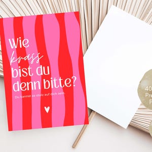 May include: A rectangular postcard with a red and pink wavy striped background. The text reads "Wie krass bist du denn bitte?" and "Du kannst so stolz auf dich sein." with a small white heart icon. A second blank white postcard is positioned next to it.