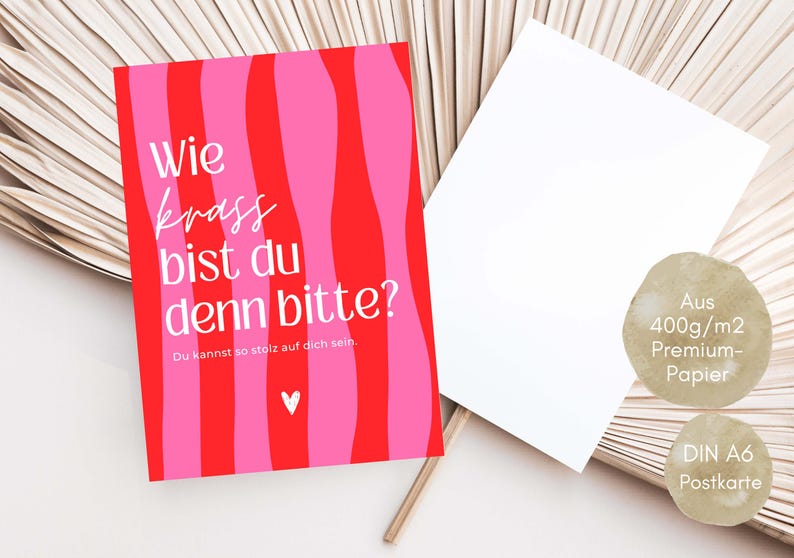 May include: A rectangular postcard with a red and pink wavy striped background. The text reads "Wie krass bist du denn bitte?" and "Du kannst so stolz auf dich sein." with a small white heart icon. A second blank white postcard is positioned next to it.