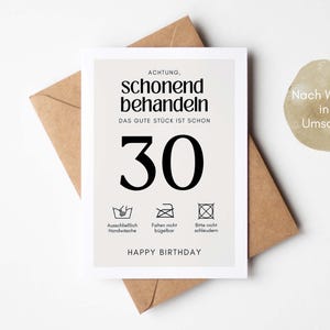 May include: A white card with black text that reads "ACHTUNG, schonend behandeln DAS GUTE STUCK IST SCHON 30" with three black and white icons below the text. The icons represent washing instructions. The text "HAPPY BIRTHDAY" is below the icons. The card is sitting on a brown envelope.