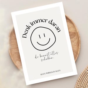 May include: A rectangular print with a light beige background features a black smiley face and the German text "Denk immer daran" and "du kannst alles schaffen." The print is on a wooden plate.