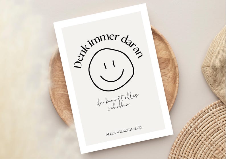 May include: A rectangular print with a light beige background features a black smiley face and the German text "Denk immer daran" and "du kannst alles schaffen." The print is on a wooden plate.