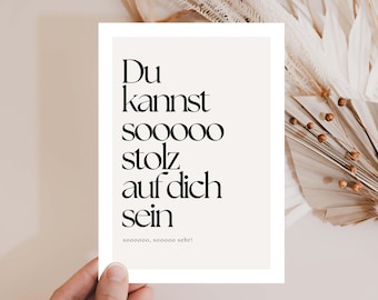 May include: A rectangular card with the German text "Du kannst sooooo stolz auf dich sein" in black, elegant font. The card is held by a hand, with dried plants in the background. The text translates to "You can be so proud of yourself."