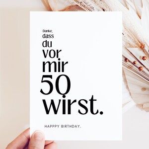 May include: A white card with black text that reads "Danke, dass du vor mir 50 wirst." and "HAPPY BIRTHDAY."  The card is held in a hand against a background of dried palm leaves.