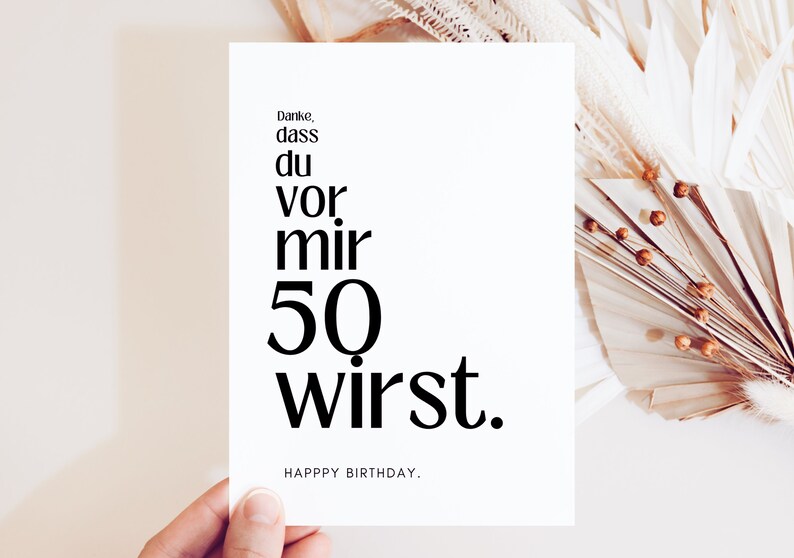 May include: A white card with black text that reads "Danke, dass du vor mir 50 wirst." and "HAPPY BIRTHDAY."  The card is held in a hand against a background of dried palm leaves.