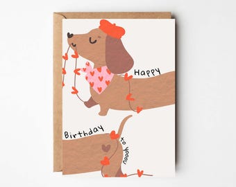 Puede incluir: Tarjeta de felicitación con un perro salchicha de dibujos animados que lleva una boina roja y una bufanda rosa con corazones. El perro sostiene una guirnalda de corazones rojos. La tarjeta dice "Feliz cumpleaños".