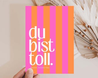 May include: A greeting card with vertical stripes in bright pink and orange. The white text reads "du bist toll." with "soooo, soooo toll" at the bottom. The card is held by a hand, with dried flowers in the background.