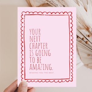 May include: A pink greeting card with a red scalloped border. The card features the text "YOUR NEXT CHAPTER IS GOING TO BE AMAZING. WISHING YOU THE BEST." The card is held by a hand.