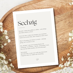 May include: A printed card with the German word "Sechzig" at the top, surrounded by text and a smiley face. The card is on a wooden tray, with white flowers around the edges.