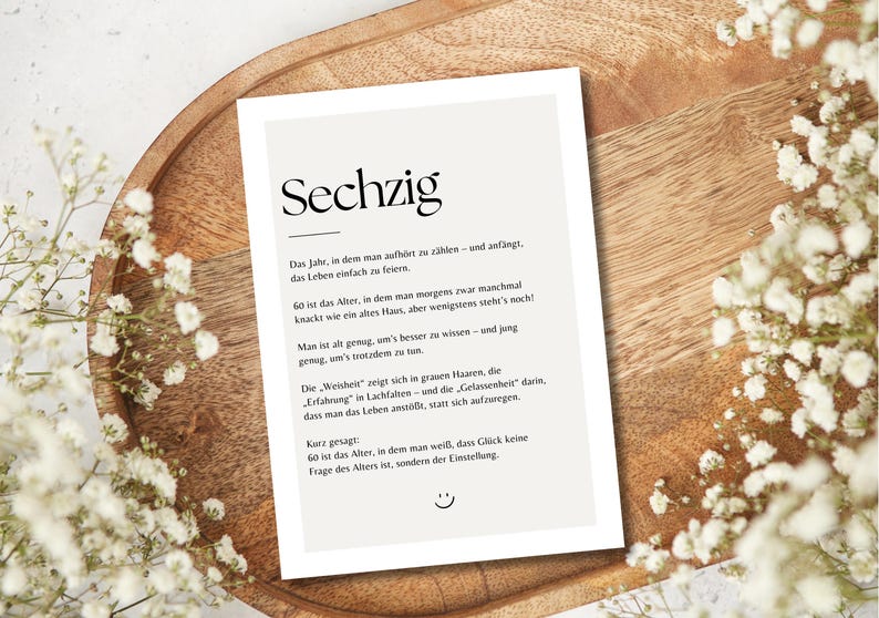 May include: A printed card with the German word "Sechzig" at the top, surrounded by text and a smiley face. The card is on a wooden tray, with white flowers around the edges.