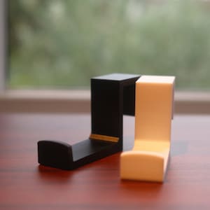 May include: Two wall hooks, one black and one white, are made of plastic and are shaped like a J. They are sitting on a wooden surface.