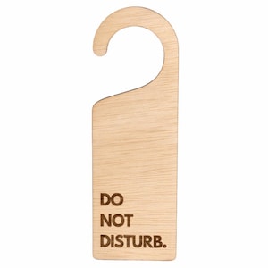May include: A light-colored wooden door hanger with a smooth grain texture. The hanger features a curved hook at the top for placement on a door handle. The text DO NOT DISTURB. is engraved in a dark brown, sans-serif font on the lower section.