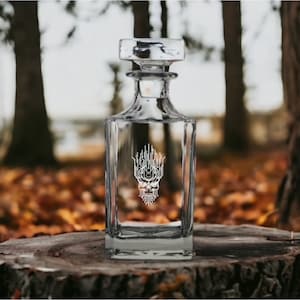 May include: A clear glass decanter with a square body and a stopper. The decanter has an etched design of a skull with a beard and a crown.